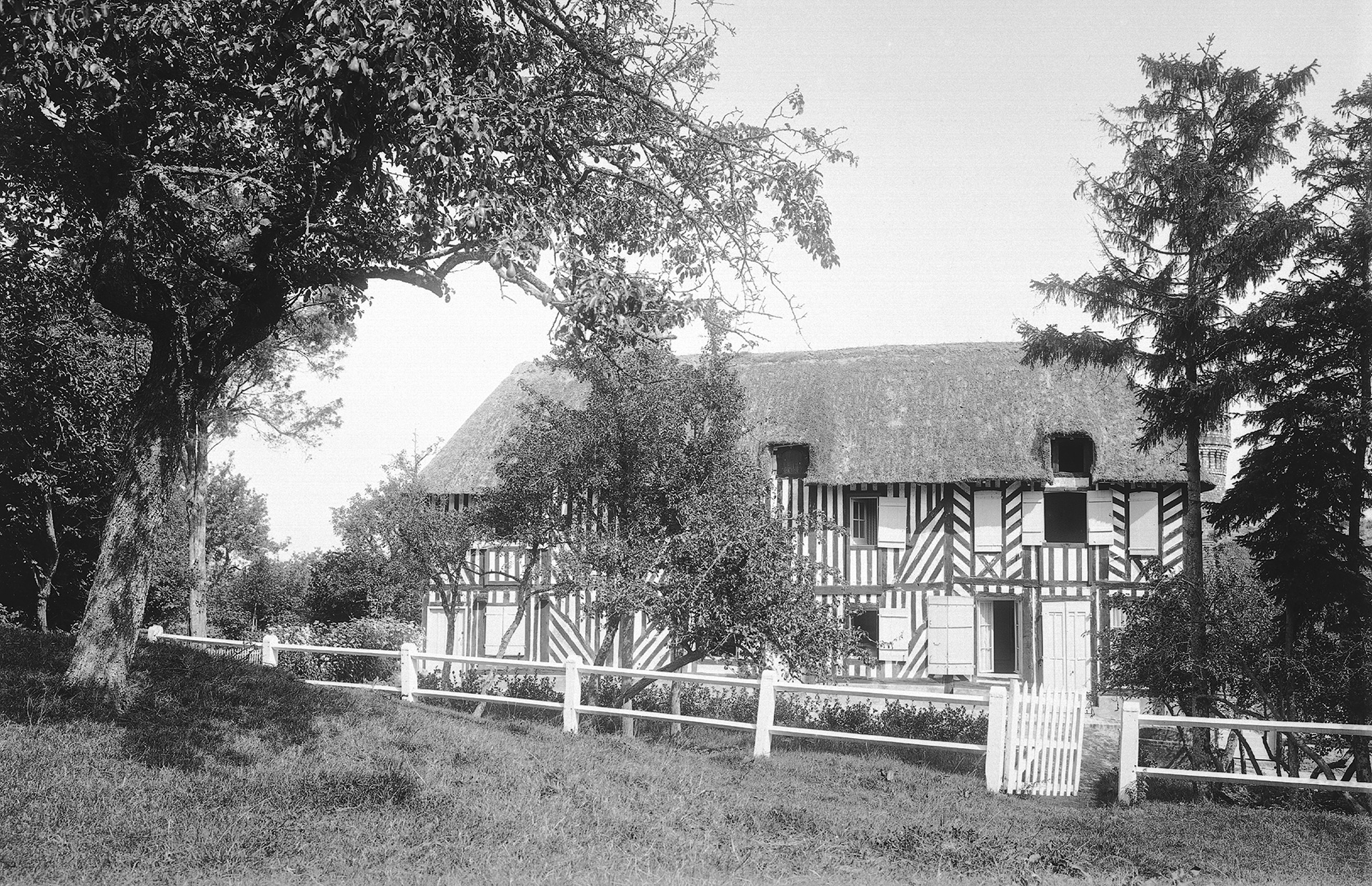 Photographie en noir et blanc. Façade d'une maison à pan de bois avec un toit en chaume dans un jardin. Au premier plan une clôture peinte en blanc.