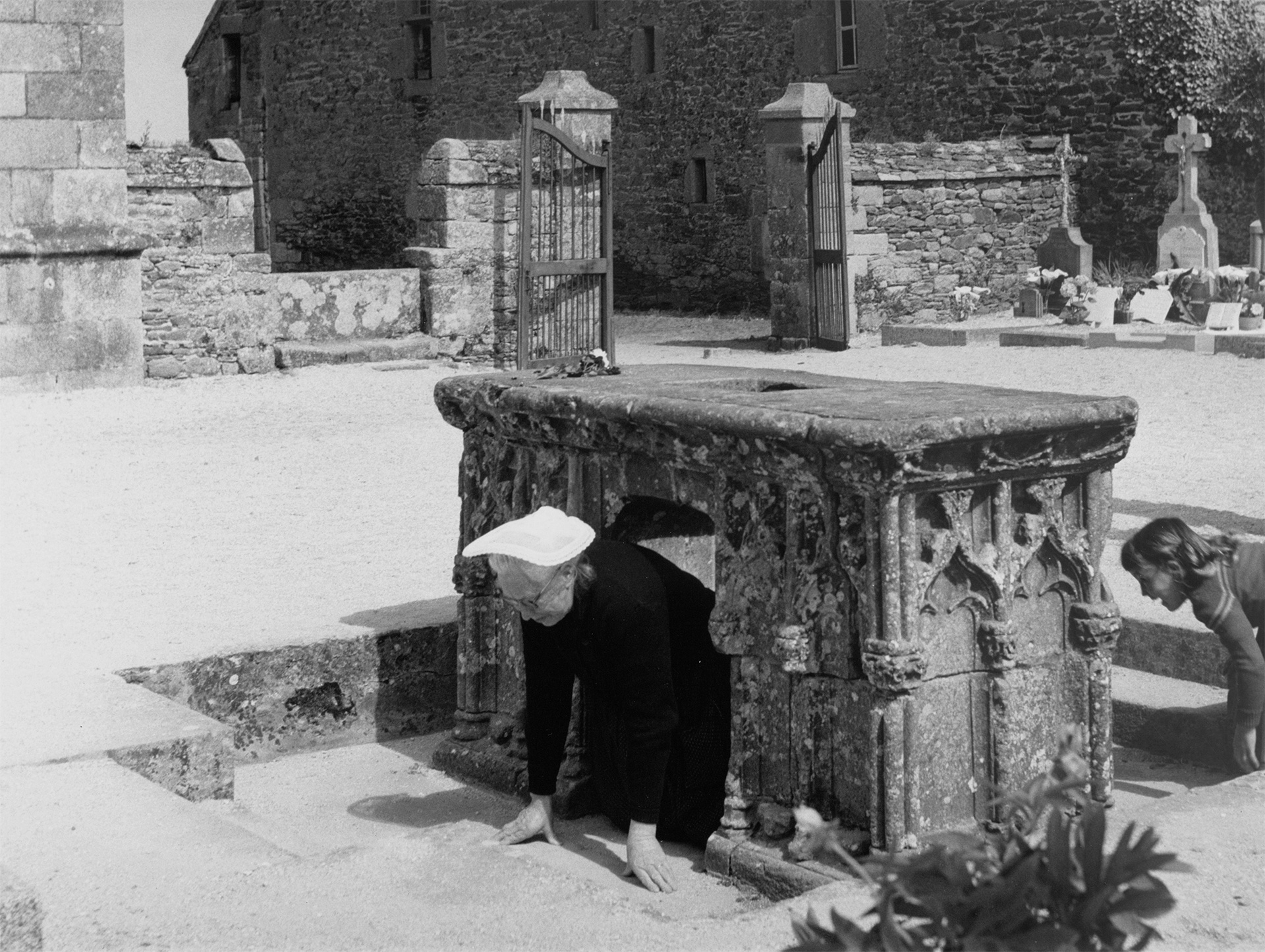 Une femme portant une coiffe bretonne passe sous en autel en pierre, cénotaphe de saint Yves, dans le cimetière de Minihy-Tréguier 