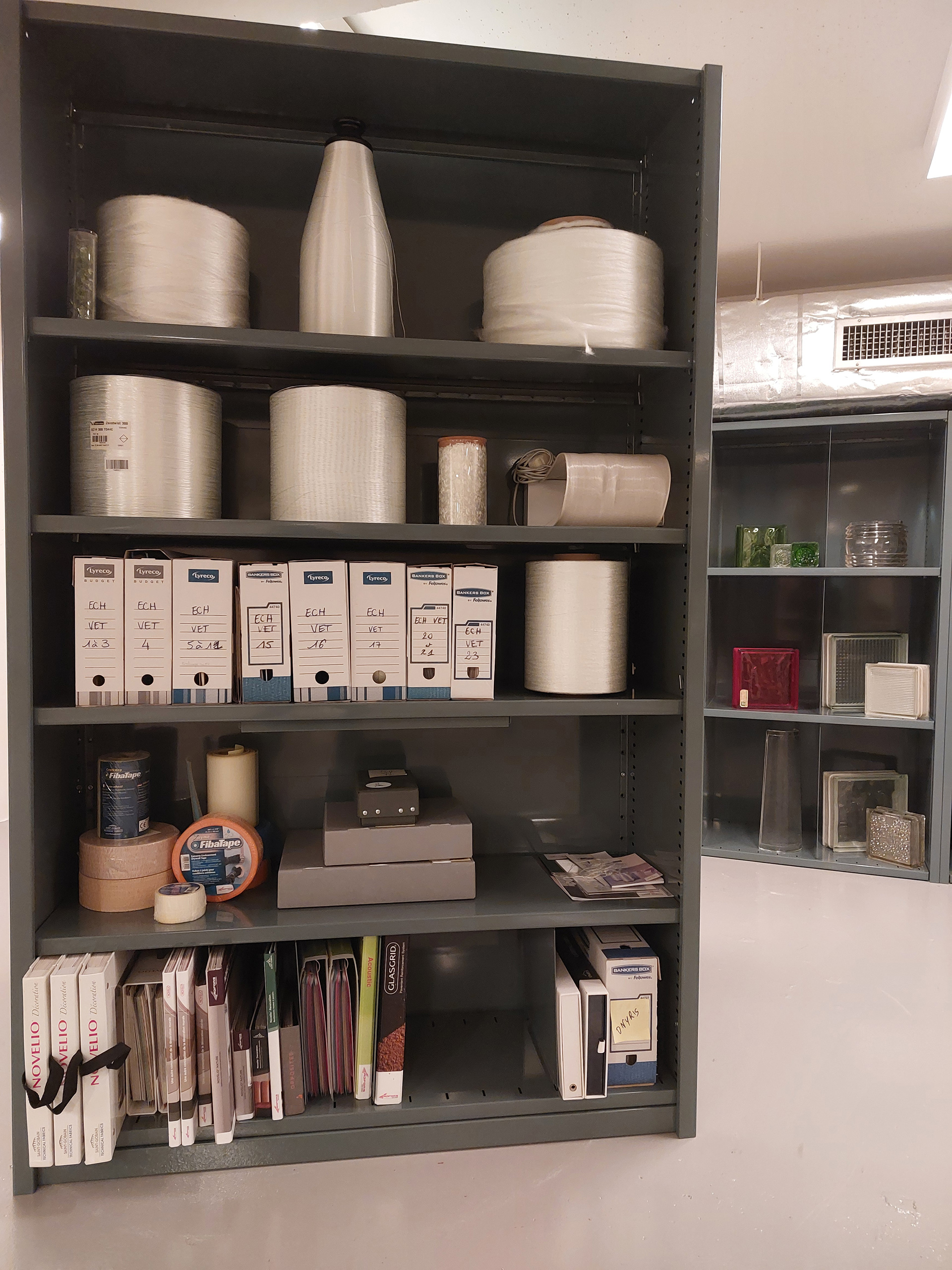 Section présentant les échantillons de l’activité renforcement : bobines de ruban et de fils de fibre de verre de marque Vetrotex, bandes FibaTape en maille de fibre de verre pour le renforcement des joints Adfors, et revêtements muraux décoratifs Novelio en fibre de verre.  En arrière-plan, ensemble de moulages en verre, XXe s.