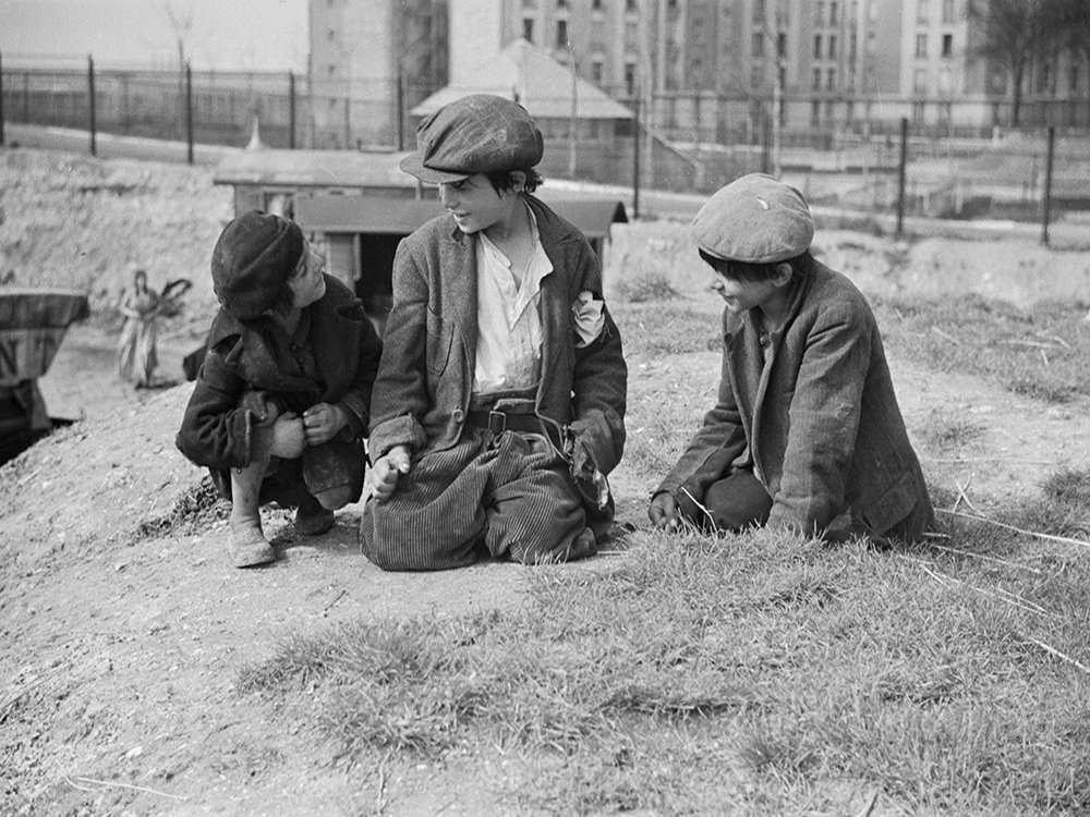 3 jeunes garçons assis par terre. En contre-bas, en arrière plan, des roulottes.