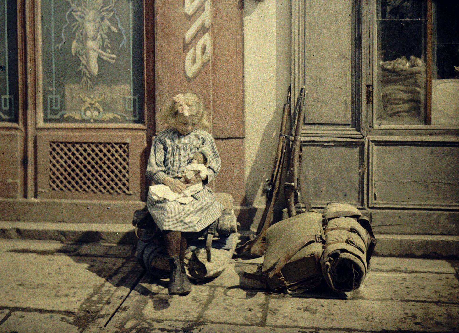 Fernand Cuville, Petite rémoise : fillette jouant à la poupée dans la rue ; À côté delle, fusils et sac de soldat, Reims, 1917