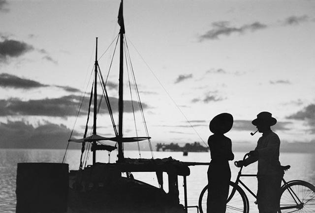 un couple en hombre chinoise devant un ponton et un bateau à voile sur fond de couché de soleil.