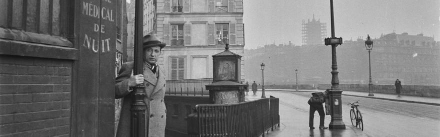 Un homme en par-dessus et chapeau regarde l'objectif dans un décor parisien.
