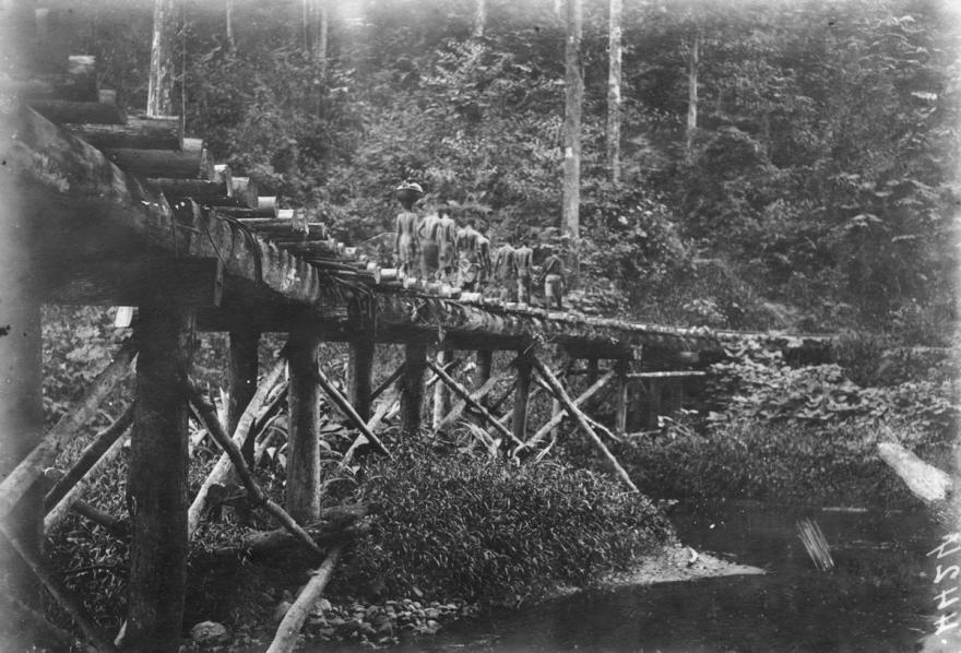 Un groupe franchit un pont ferroviaire sur la Petite Malumé : un tirailleur précède six homme et une femme portant une charge sur sa tête.