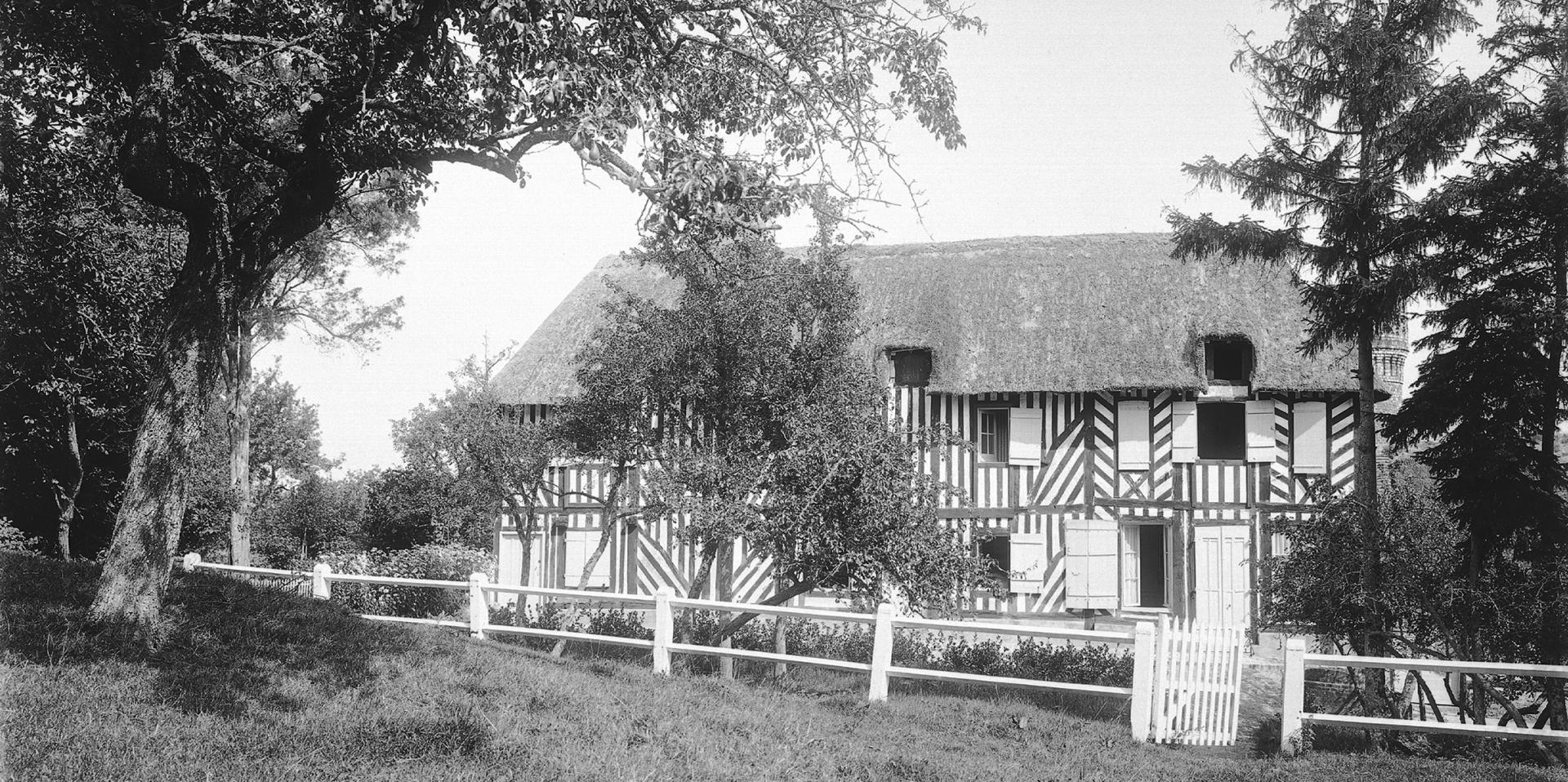Photographie en noir et blanc. Façade d'une maison à pan de bois avec un toit en chaume dans un jardin. Au premier plan une clôture peinte en blanc.